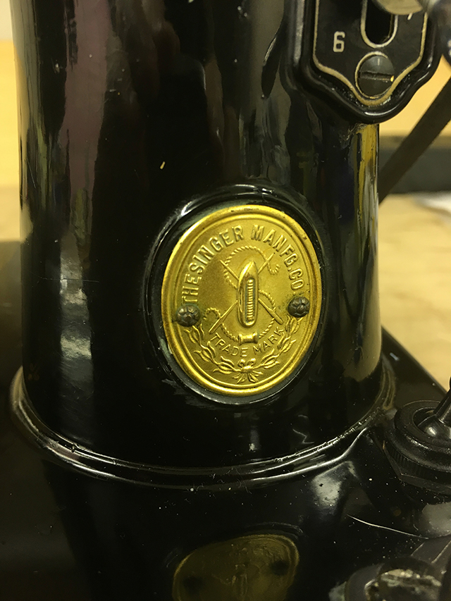 A brass seal is held onto the sewing machine pillar with two ornate rivets. The seal reads "THE SINGER MANFG. CO. / TRADEMARK," The seal shows an old fashoned vibrating shuttle&ndash;style bobbin with two machine needles crossed behind it. The letter 'S' is formed by a length of thread.